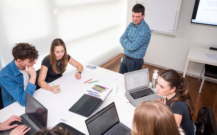 Studenten zitten rond een tafel. Sommigen hebben een laptop voor zich. Een meisje is aan het tekenen, terwijl een staande docent toekijkt. 