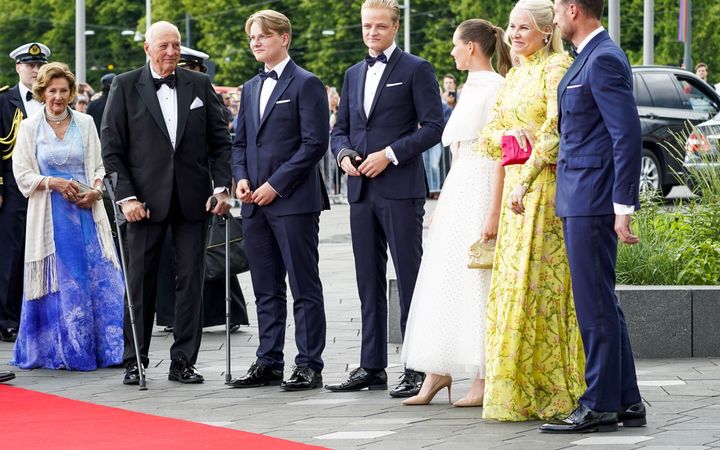 Zeven mensen staan achter een rode loper in galakleding.