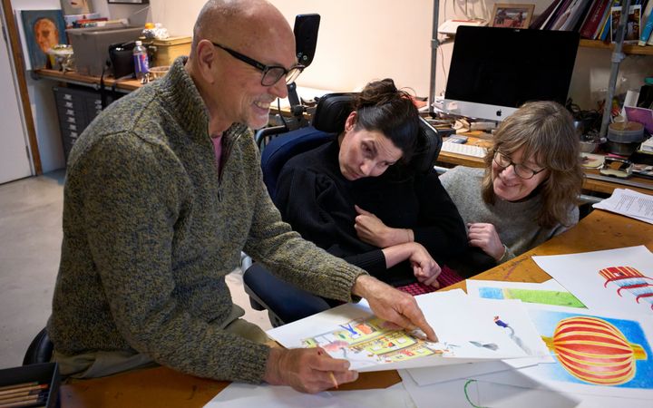 Drie mensen kijken naar kleurrijke illustraties op grote witte vellen. Een man met een snor en bril wijst iets aan. Twee vrouwen kijken toe, de ene zit in een rolstoel. 