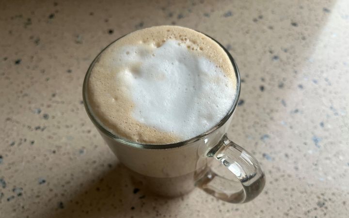 Een glas  op een licht-granieten aanrecht. In het glas zit schuim dat grotendeels wit is, en aan de randen lichtbruin. Onderin het glas zit bruine vloeistof.