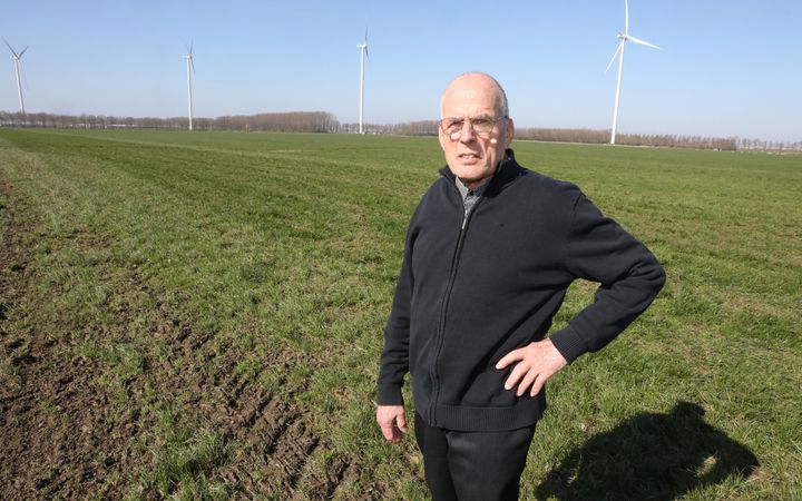 Een ernstig kijkende, kale man in zwarte kleding staat in een groen weiland. Op de achtergrond zijn windmolens te zien.