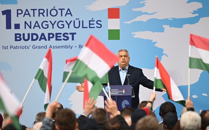 Een man in een donkerblauw jasje met lichtblauw overhemd spreekt mensen toe vanachter een katheder. Ze zwaaien met rood-wit-groene vlaggetjes.