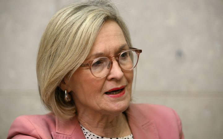 Een pratende vrouw van rond de zestig met geblondeerd haar, een bruine bril en roodgestifte lippen, gekleed in een roze jasje. 