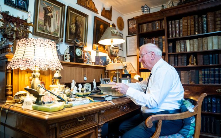 Een oudere man in een wit overhemd achter een bureau, met om zich heen veel kunstvoorwerpen.