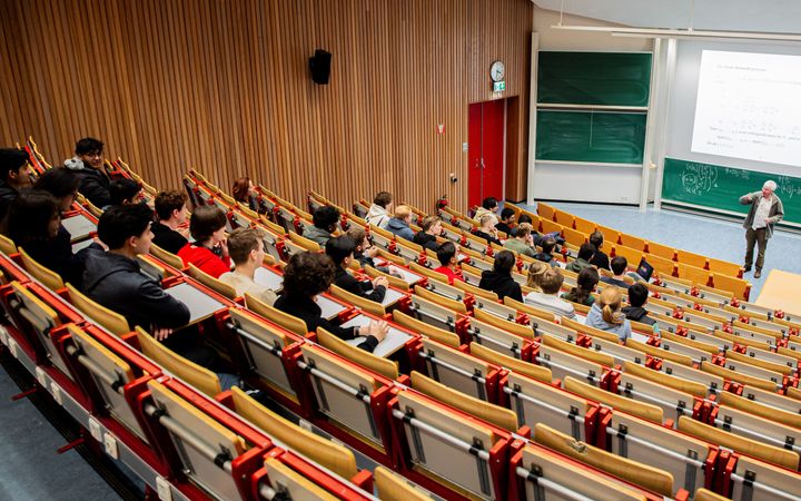 Studenten zitten in een collegezaal. Voorin staat een lector die uitleg geeft. Op het bord achter hem staan formules geprojecteerd.