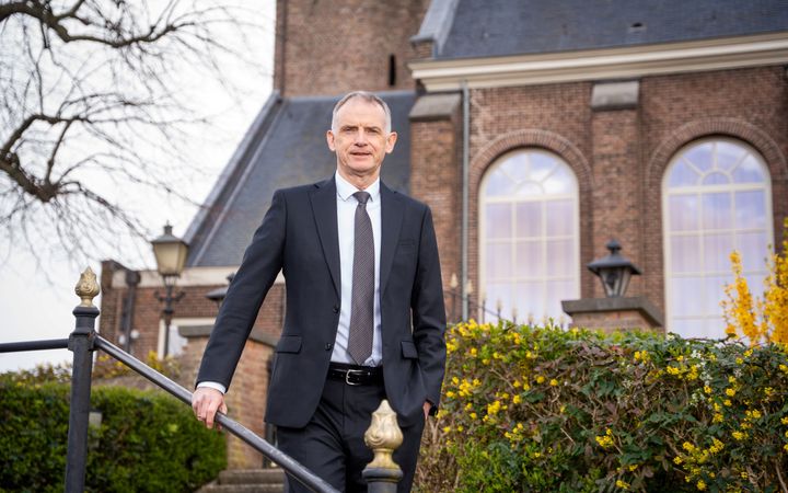 Man in een donker pak met een stropdas staat voor een kerkgebouw met grote ramen. 