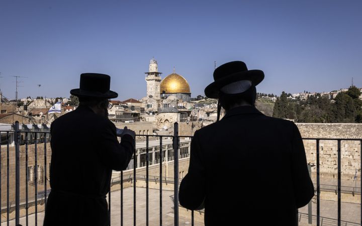 Twee in het zwart geklede mannen met zwarte hoeden staan voor een zwart hek. Daarachter ligt de oude stad van Jeruzalem.