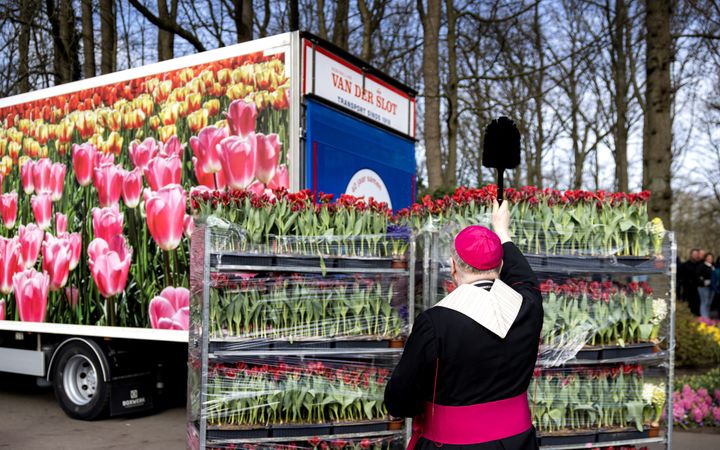 Een man in zwart-paars gewaad houdt een wijwaterkwast boven een kar bloemen en spreekt een zegen uit.