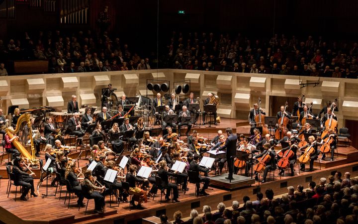 In een grote concertzaal zit een orkest te spelen. Op de bok staat een dirigent.