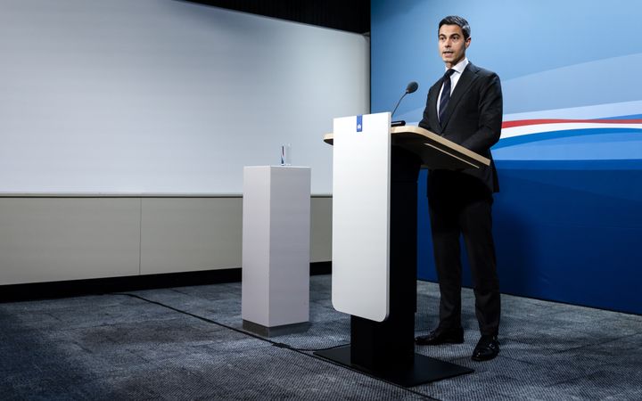 man in donker pak achter spreekgestoelte voor een blauw decor met rood-wit-blauwe sjerp