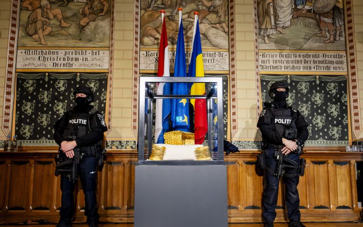 Twee mannen in donkere kleding, helm op, bivakmuts op, geweer vast, staan naast een sokkel met in een vitrine drie gouden voorwerpen.