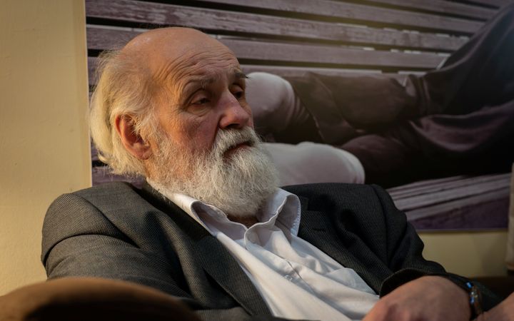 oude man met wit haar en witte baard in grijs jasje over een wit overhemd zit op een bank en kijkt droevig.