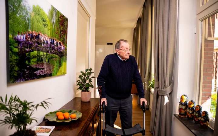 Een oude man staat achter zijn rollator, in een gang waar links aan de muur een grote familiefoto hangt; rechts staan in de vensterbank vier ma&shy;troesj&shy;ka&rsquo;s.