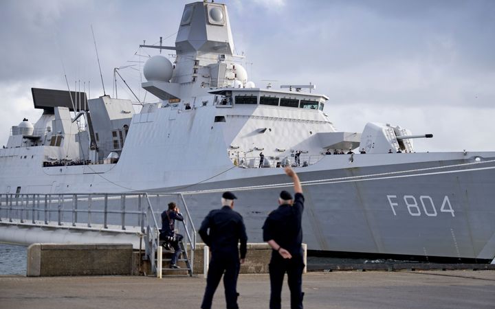 Een groot, grijs marinefregat vertrekt uit een haven. Twee mannen zwaaien naar het schip.  