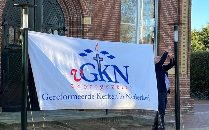 Een grote vlag met als opschrift ”voortgezette Gereformeerde Kerken in Nederland” wordt gespannen tussen twee palen voor een kerkgebouw.