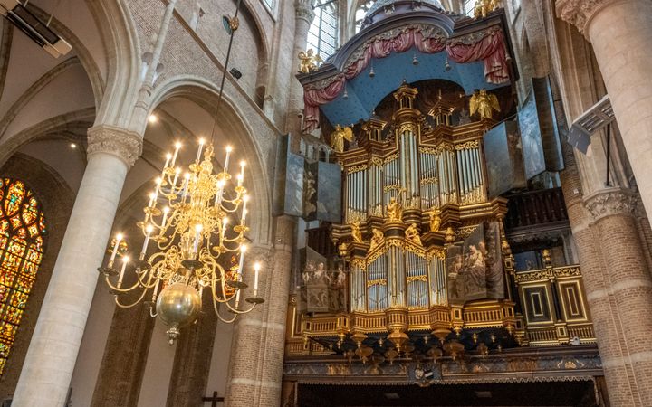 In een eeuwenoude kerk hangt een groot kerkorgel. Het orgel telt veel met goudverf beschilderde beelden. Aan weerszijden van de orgelkas zijn luiken aangebracht. Voor het orgel hangt een brandende kroonluchter.