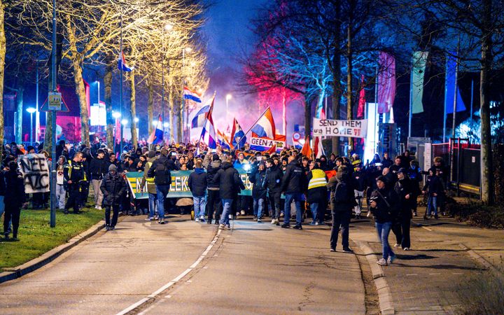 Honderden mensen lopen midden op straat en houden rood-wit-blauwe vlaggen omhoog en borden met ”Send them home”. 