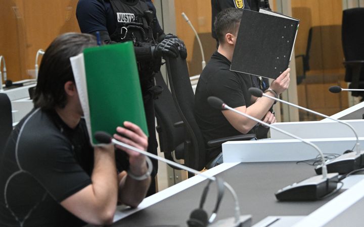 Twee mannen zitten in een rechtbank en houden boeken voor hun gezicht. 