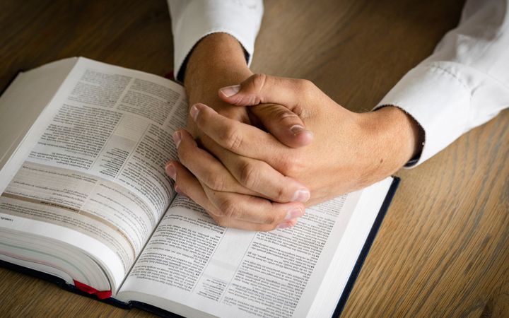 Gevouwen handen van iemand die een wit overhemd draagt, boven een opengeslagen Bijbel.