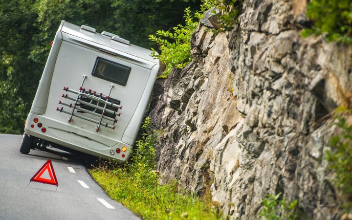 Een witte camper staat naast de weg, schuin tegen een rotswand. Op de voorgrond een gevarendriehoek. Op de achtergrond wat bos en groene struiken.