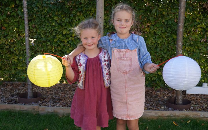 Twee kinderen staan in een tuin met de armen om elkaar heen geslagen. Ze houden allebei een lampion vast.