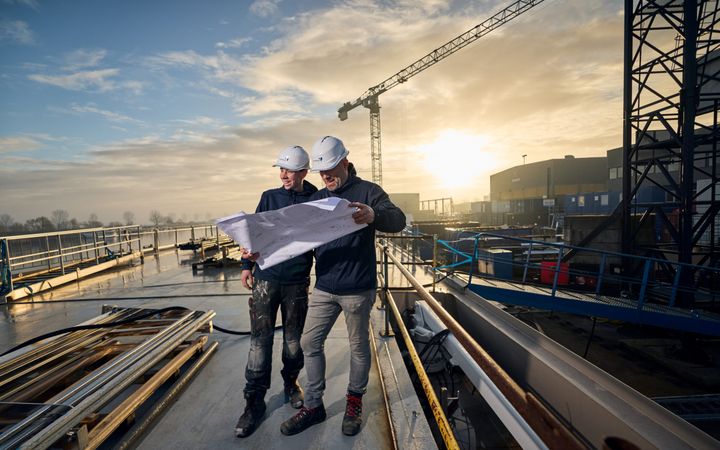 Twee monteurs met helm lezen een werkplan aan boord van een schip. 