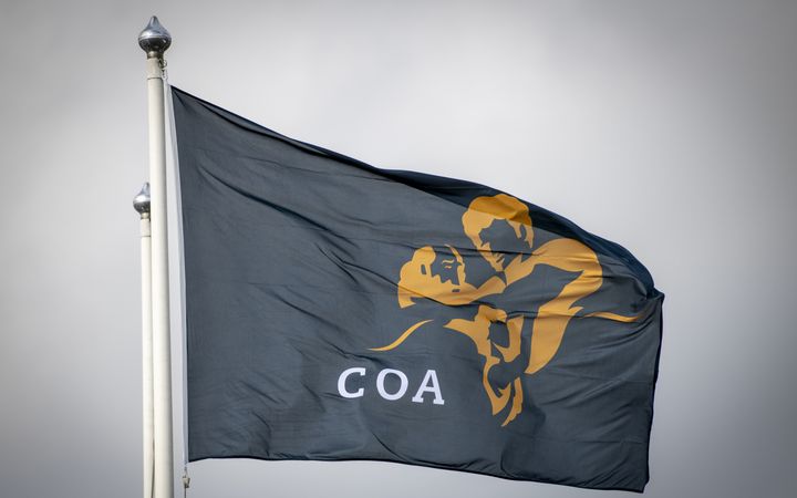 Een donkerblauwe vlag met daarop in geel het logo van Centraal Orgaan opvang asielzoekers met in witte letters de afkorting COA wappert in de wind.