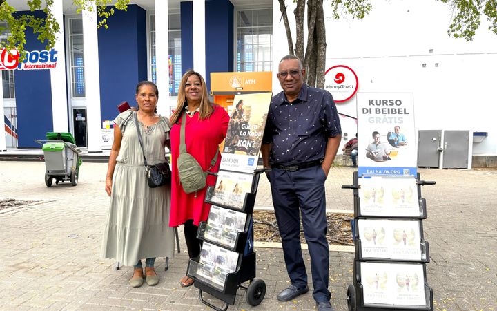 Een man met zijn handen in de zak en twee vrouwen staan  op een plein met karretjes waarop gratis Bijbelcursussen worden aangeboden.