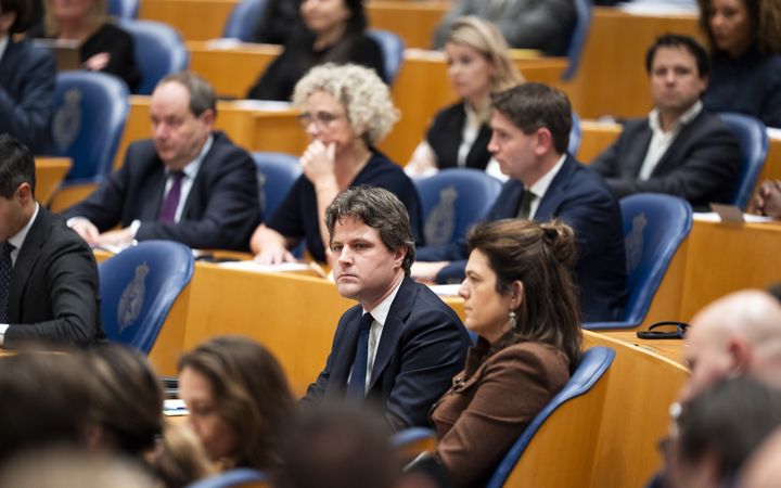 Mannen en vrouwen zitten op blauwe stoelen in het Tweede Kamergebouw. Ze kijken gespannen. 