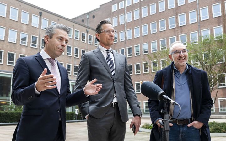 Drie mannen, twee in pak met das, staan buiten voor een microfoon; achter hen is een hoog gebouw te zien met talloze ramen.