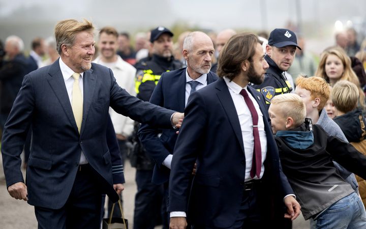 De koning begroet omstanders. Om hem heen lopen beveiligers en politieagenten.