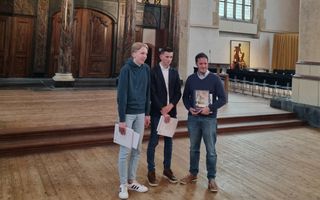 Drie organisten staan lachend voor een oud orgel in een kerk.