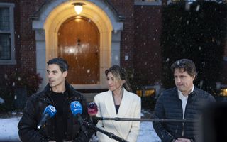 Drie personen met voor hen microfoons en achter hen de monumentale toegangsdeur met halve ronde boog. Ondertussen dwarrelen de sneeuwvlokken neer.