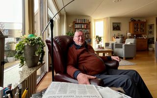 Oudere man met roodbruin vest zit in een roodleren leunstoel, onder een staande lamp, en lacht in de camera.