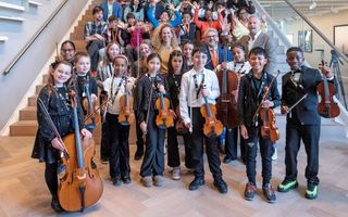 Kinderen kijken blij in de camera. Ze hebben een viool of een cello vast. Achter hen zitten of staan volwassenen op een trap.