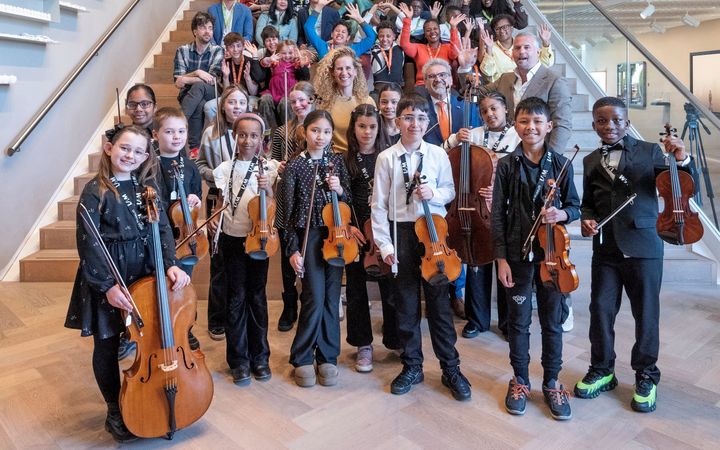 Kinderen kijken blij in de camera. Ze hebben een viool of een cello vast. Achter hen zitten of staan volwassenen op een trap.