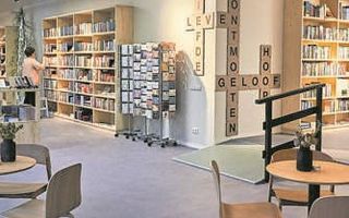 Interieur met boekenkasten. Een vrouw sorteert ze. Op de voorgrond tafeltjes en stoelen.