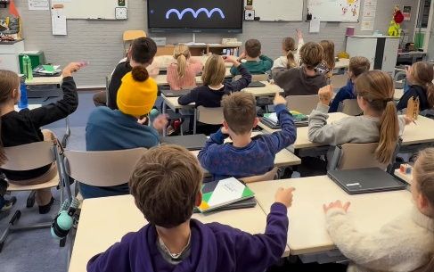Kinderen in klas volgen met hun vinger een golvend patroon dat op het digitale bord voor in de klas staat.