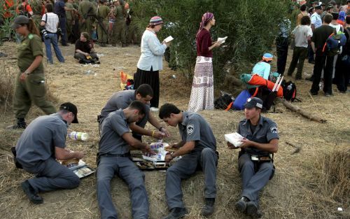 KFAR MAIMON â€“ Joodse vrouwen bidden tijdens een protestactie, terwijl agenten van de IsraÃ«lische oproerpolitie het avondeten gebruiken. Duizenden actievoerders wachtten gisteren bij het dorpje Kfar Maimon op de Westelijke Jordaanoever tot ze hun mars n