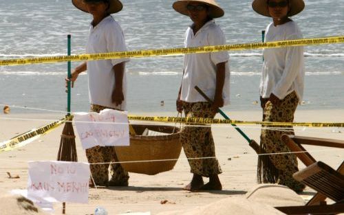 JIMBARAN â€“ Balinese strandschoonmakers passeren de plaats waar afgelopen zaterdag een bom ontplofte. De Australische regering heeft toeristen ertoe opgeroepen vakantiekiekjes ter beschikking te stellen voor het politieonderzoek. Volgens president Yudhoy