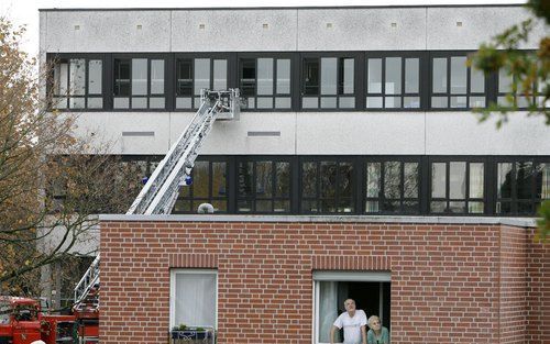 EMSDETTEN - Twee buren kijken uit een raam naast het schoolgebouw in Emsdetten waar een gijzeling plaatsvond. In de achtergrond is het schoolgebouw te zien waar de vensters open staan en er een ladderwagen tegen de gevel staat.