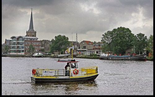 Een klein geel zwart geschilderd pontje dobbert midden op de rivier. Het is de levensader tussen Ouderkerk en Nieuwerkerk.