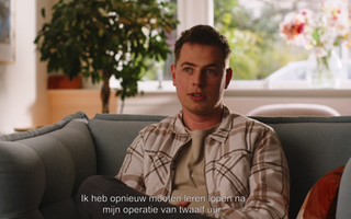 Een jongen in een geruite blouse zit op een bank. Achter hem een plant op stam.