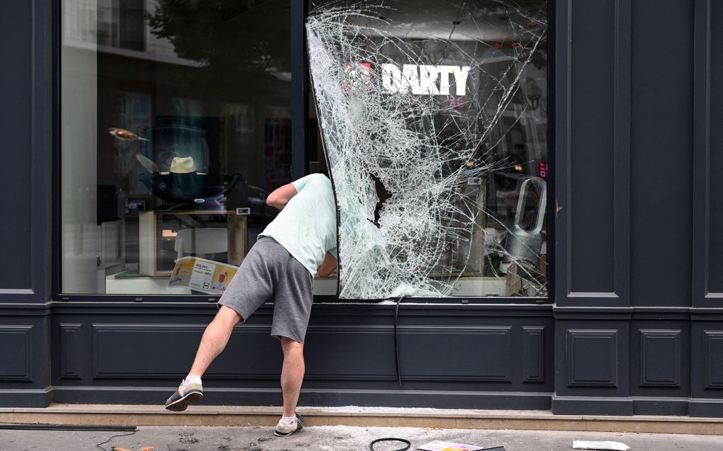 Een man kijkt door een gat van een vernielde winkelruit. Menig winkel moet het ontgelden tijdens de rellen in Frankrijk. beeld AFP