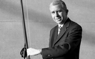 Een man in een donker pak houdt glimlachend een snaarinstrument met een lange hals vast.