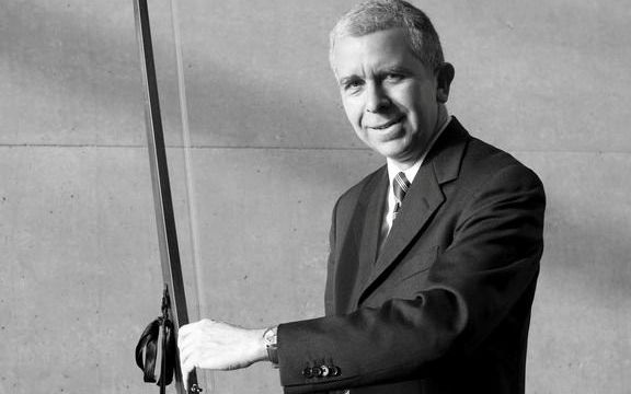 Een man in een donker pak houdt glimlachend een snaarinstrument met een lange hals vast.