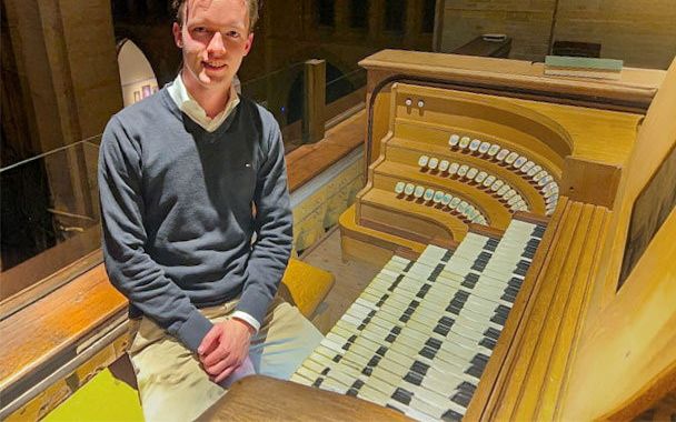 Een jongeman in de grijze trui zit op de orgelbank naast de vier klavieren van een groot orgel.