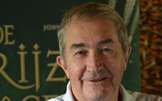 John Flanagan, met witte polo, op de voorgrond. Achter hem een poster van een van zijn boeken.