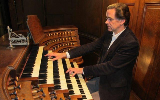 Een man, gekleed in een zwart colbert, zit achter de klavieren van een kerkorgel. Aan beide zijden van de vijf klavieren van dit instrument zitten rijen registers.