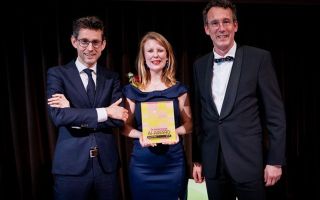 Een vrouw met donkerblauwe galajurk houdt een plaquette vast waarop staat: De Nederlandse AI-Award. Links van haar staat een man met een donkerblauw pak en een stropdas, rechts van haar staat een man met een antraciet pak en een strikje. 